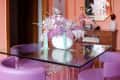 Glass dining table with purple chairs, floral centerpiece, and pink walls in a modern room.