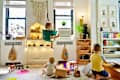 Children playing in a colorful playroom with toys, a hanging swing, and a dollhouse under a macrame wall hanging.