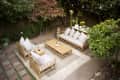 Outdoor patio with wooden furniture, gray cushions, string lights, and potted plants surrounded by lush greenery.