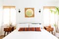 Bedroom with white bedding, colorful striped pillow, wooden nightstands, wall art, and potted plant by the window.