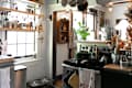 Cozy kitchen with open shelves displaying bottles, pots hanging above, and a green stove with utensils and plants.