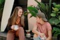Two people sitting on a wooden deck surrounded by lush plants, holding mugs and smiling at each other.