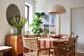 Dining room with woven chairs, wooden table, striped runner, large plant, round mirror, and pendant light.