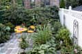 Small garden patio with yellow table and chairs, surrounded by lush greenery and a white fence.