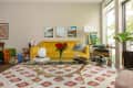 Living room with yellow sofa, glass coffee table, red flowers, and wall art, featuring large windows and a patterned rug.