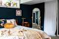 Bedroom with dark blue walls, a bed with colorful pillows, a gold round cushion, and a tall mirror beside a window.