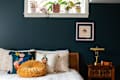 Bedroom with dark teal walls, decorative pillows on bed, brass lamp on wooden nightstand, and plants on window ledge.
