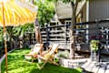 Two wooden deck chairs under a yellow striped umbrella on a grassy patio with trees and a black railing.