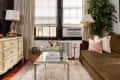 Cozy living room with olive sofa, glass coffee table, floral arrangement, and cream dresser under soft lighting.
