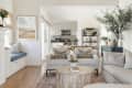 Open-concept living room with neutral sofas, round wooden coffee table, and view into a white kitchen with open shelving.