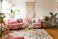 Cozy living room with pink sectional sofa, colorful pillows, macramé wall hanging, plants, and a glass coffee table.