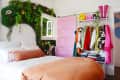 Bedroom with pink door, lush green wall decor, colorful clothes rack, and a bed with orange bedding.