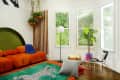 Orange tufted sofa, green rug with leopard design, hanging plants, and a floor lamp in a bright room with large windows.