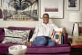 Woman sitting on a purple sofa with colorful cushions, framed art, and a lamp in the background.