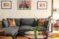 Cozy living room with a gray sectional sofa, orange floral pillows, glass coffee table, and colorful framed artwork on the walls.