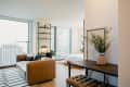 Modern apartment with brown sofa, black and white rug, wall shelves, and a desk with a plant, overlooking cityscape.