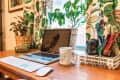 Home office desk with laptop, keyboard, mouse, coffee mug, books, and plants near colorful patterned curtains.
