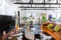 Bright living room with orange sofa, colorful pillows, glass coffee table, plants, and exposed beams.