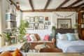 Cozy living room with a tan sofa, colorful pillows, plants, bookshelves, and a gallery wall under exposed wooden beams.