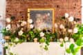 Floral arrangement with white roses and greenery on a mantel, framed painting on a brick wall backdrop.