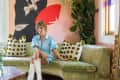Woman in blue dress sitting on green velvet sofa with polka dot pillows, large plant, and abstract wall art in living room.