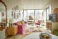 Bright living room with pink sofa, colorful rug, gallery wall, and large windows. Mid-century furniture and TV on dresser.