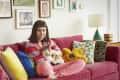 Woman sitting on a red sofa with colorful pillows, holding a cat, surrounded by framed art on the wall.