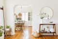 Entryway with a round mirror, console table, and vase of flowers, leading to a dining room with wicker chairs and wooden floor.