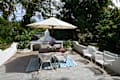 Outdoor patio with white wicker chairs, wooden table, large umbrella, and a blue patterned rug surrounded by lush greenery.