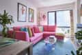 Bright living room with a pink sectional sofa, colorful cushions, glass coffee table, and stacked books by a large window.