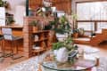 Open-plan living room with wooden staircase, glass coffee table, plants, and kitchen with bar stools and brick partition.