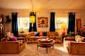 Cozy living room with patterned sofas, colorful cushions, round coffee table, vibrant artwork, and large windows with dark curtains.