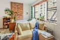 Cozy bedroom with exposed brick wall, beige sofa, potted plants, bookshelf, and large windows overlooking city buildings.