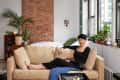 Woman in black outfit lounging on beige sofa in a cozy living room with plants, brick wall, and large windows.