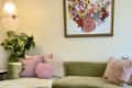 Green velvet sofa with pink cushions, floral artwork, hanging lamp, and potted plant in a cozy living room.