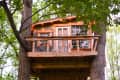 Treehouse with wooden deck, surrounded by green leaves, featuring a small table and chairs.