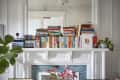 Books stacked on a white mantel with framed photos, plants, and a drawing of a cocktail.