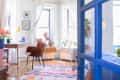 Bright living room with colorful rug, brown chair, wooden desk, and plants by large windows, viewed through blue door.
