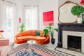 Bright living room with orange velvet sofa, pink armchair, large mirror, plants, and abstract art above a black fireplace.