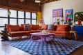 Loft living room with orange sectional sofa, colorful abstract art, purple coffee table, and blue checkered rug.