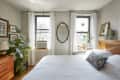 Bedroom with white bedding, wooden headboard, plants on windowsill, and framed art on walls.