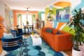 Colorful living room with orange sofa, blue chairs, abstract wall art, and plants, leading to dining area with glass doors.