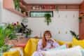 Woman in a cozy living room with yellow sofa, pink walls, and lush green plants.