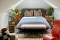 Attic bedroom with skylights, a gray bed, patterned pillows, wooden side tables, plants, and a red chair.