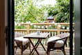 Wooden balcony with wicker chairs, a small table, string lights, and lush green trees in the background.