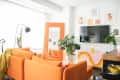 Bright living room with orange sofa, large mirror, plants, wall art, and a mounted TV.