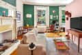 Living room with green and pink walls, beige sofa, wooden coffee table, desk, and large windows.