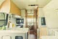 Camper interior with kitchen, sink, microwave, and dining area. Wooden cabinets, patterned wall, and view into bedroom.