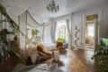 Elegant bedroom with canopy bed, chandelier, plants, and unique book tower near a large window.