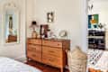 Bedroom with wooden dresser, decorative mirror, and wicker basket. View into music room with piano and painting.